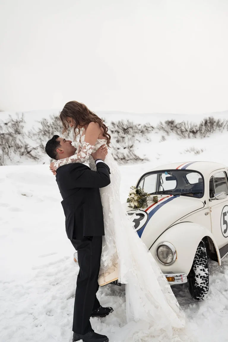 Martin lifting Carla in the snow beside a vintage Beetle during their winter elopement in Corvara